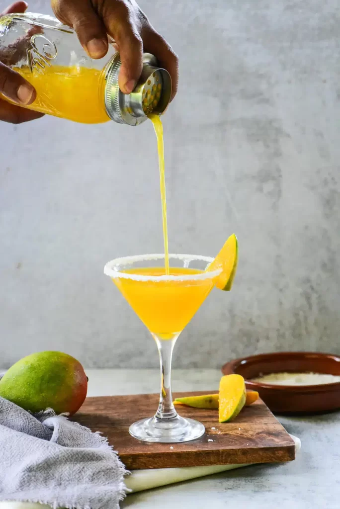 pouring mango martini in a glass placed on a wooden tray with lemon wedges