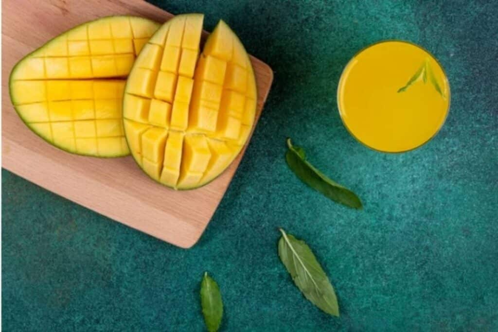 Sliced ripe mango on a wooden board with a glass of mango juice and fresh green leaves on a teal surface