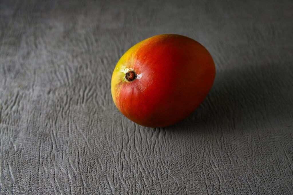 A close up of a ripe mango with a red and yellow skin resting on a textured grey surface A close up of a ripe mango with a red and yellow skin resting on a textured grey surface