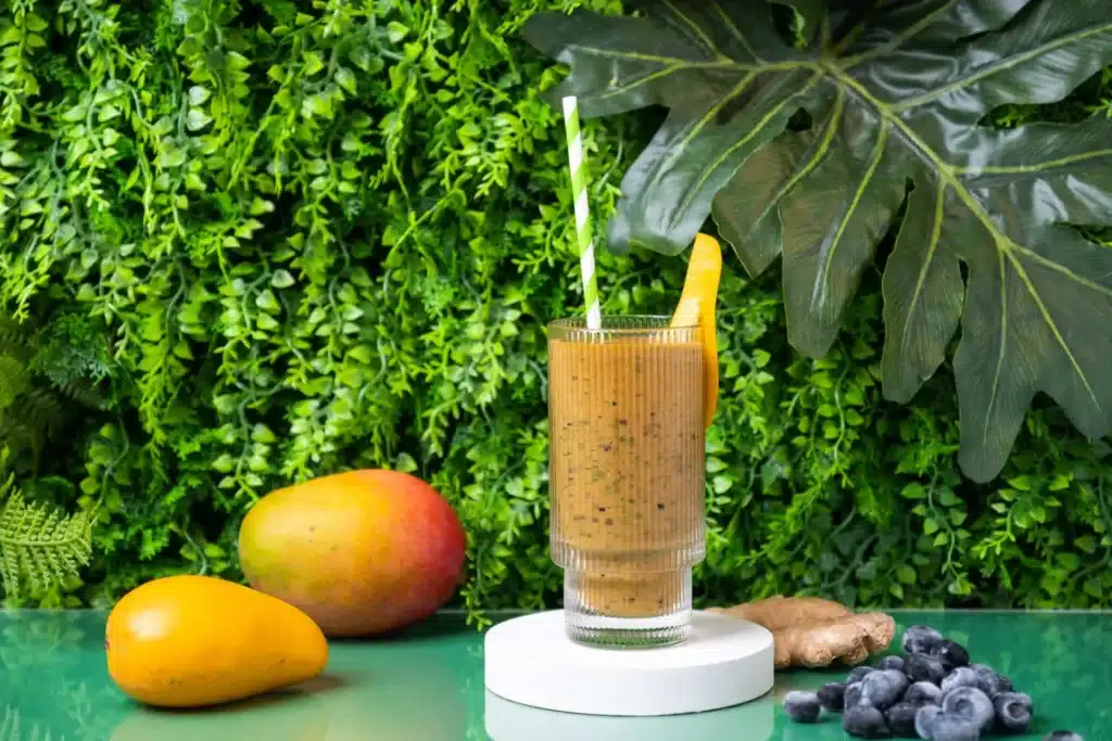 A green mango smoothie in a glass with a slice of mango surrounded by fresh mangoes and blueberries A green mango smoothie in a glass with a slice of mango surrounded by fresh mangoes and blueberries