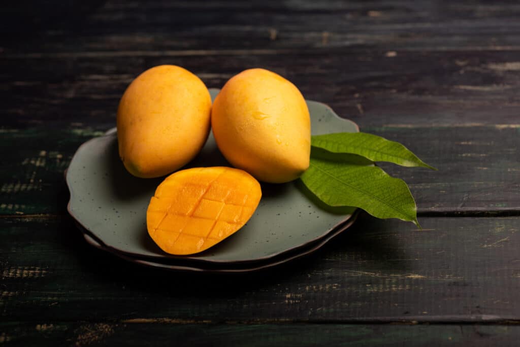 A plate with two whole mangoes and a sliced mango displaying the flesh with a crisscross pattern surrounded by green mango leaves A plate with two whole mangoes and a sliced mango displaying the flesh with a crisscross pattern surrounded by green mango leaves